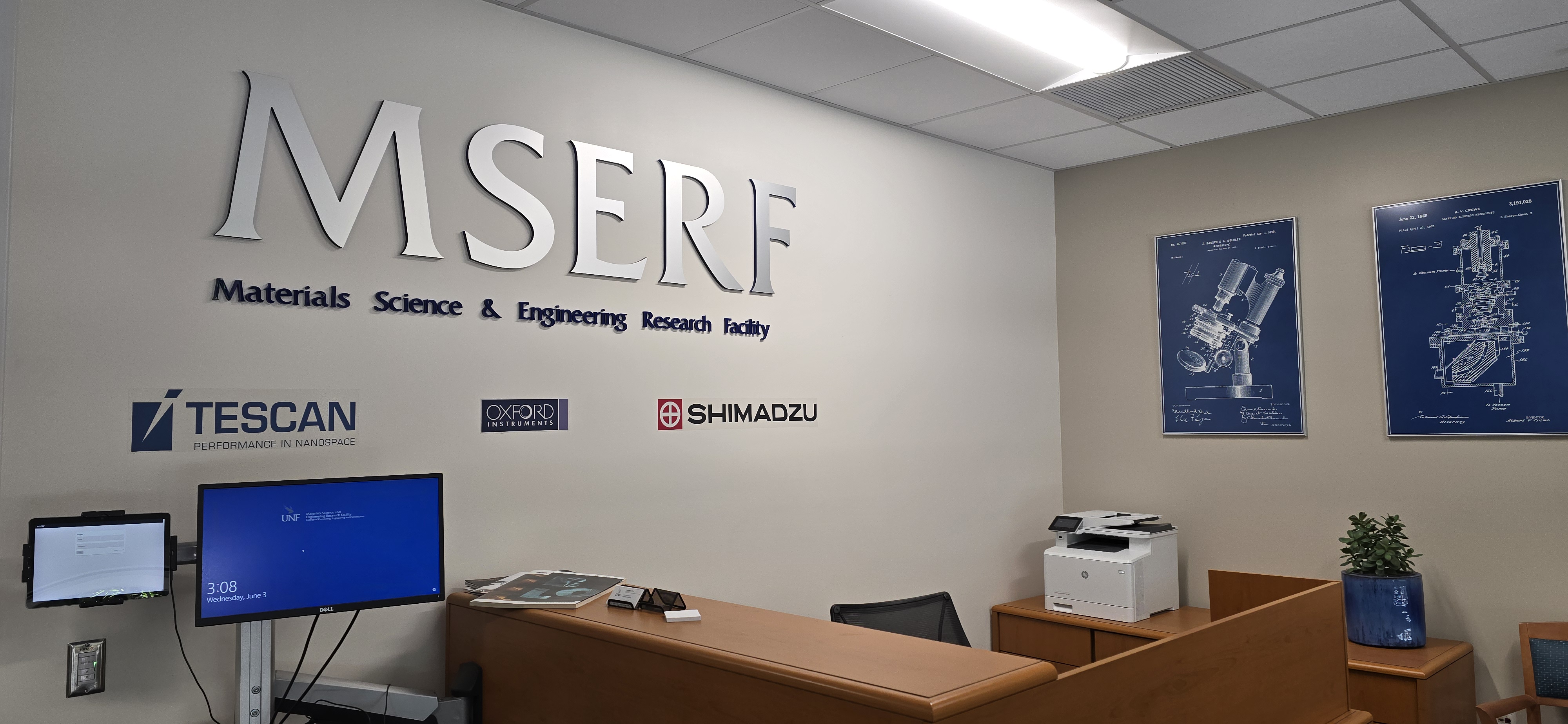 Image of the foyer as you walk into MSERF.  MSERF logo on the wall, Partner companies listed below, images of research equipment on the adjacent wall all behind a desk 