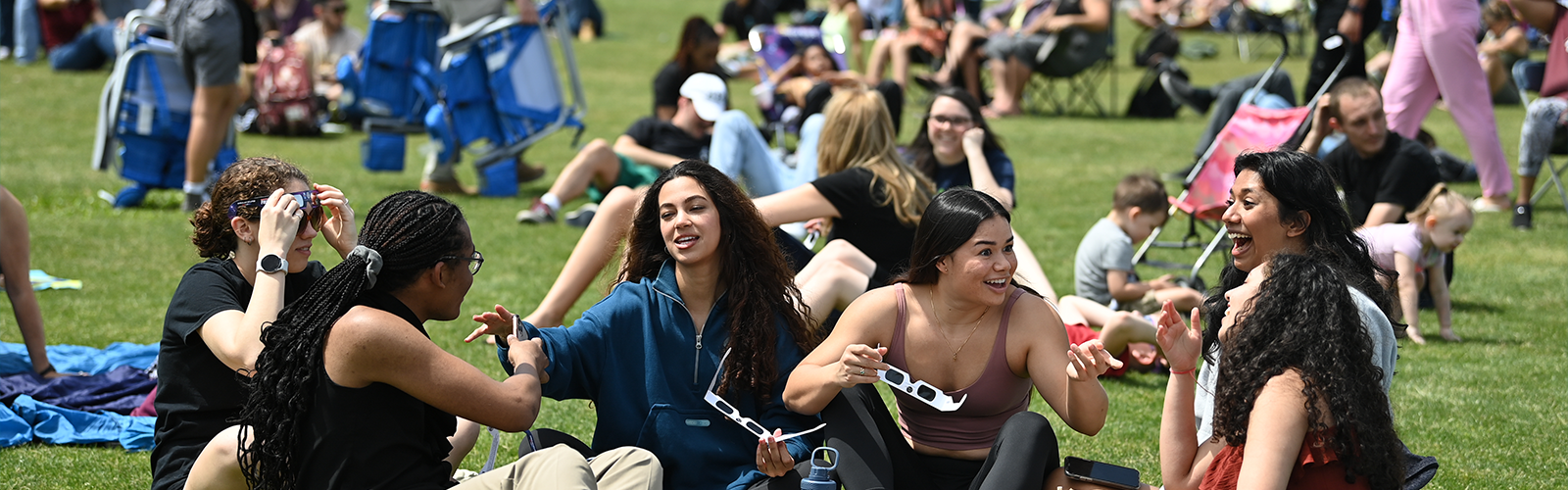 Group of students and the community sitting on grass with solar eclipse glasses socializing