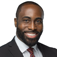 Courtney has a beard and wearing a black suit, he is smiling.