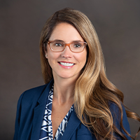 Jennifer has long hair, smiling, and wearing glasses, blue suit jacket, and blue and white blouse.