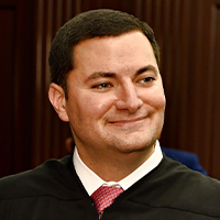 Judge Michael smiling and wearing judge robes with a white undershirt and red tie