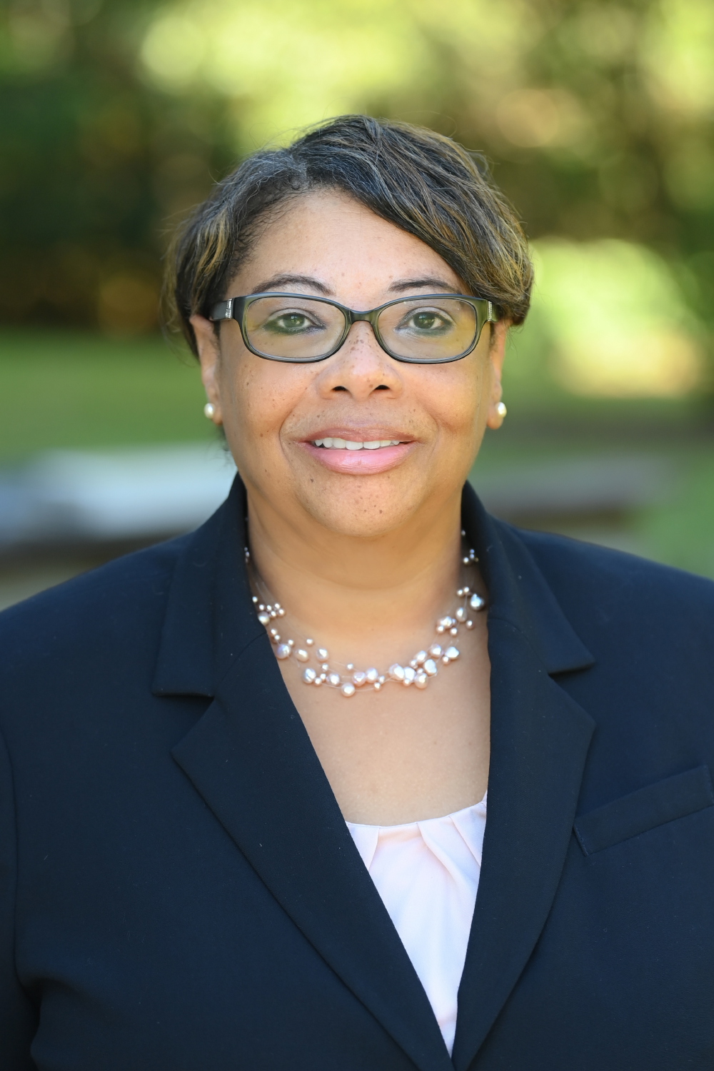 Sheila smiling and wearing glasses, navy blue suit coat over a white top, and white pearl necklace