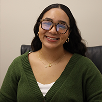 Alma smiling and wearing a glasses, two necklaces, white under shirt, and green sweater