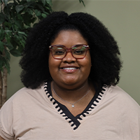 Mahalia smiling and wearing a green top with black collar, silver necklace, and glasses.