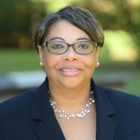 Sheila smiling and wearing glasses, navy blue suit coat over a white top, and white pearl necklace