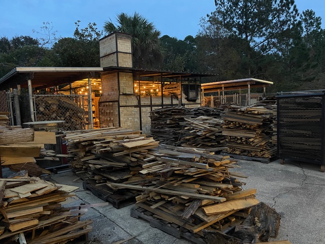 Wood yard with stacked wood piles and a structure illuminated by warm lights.