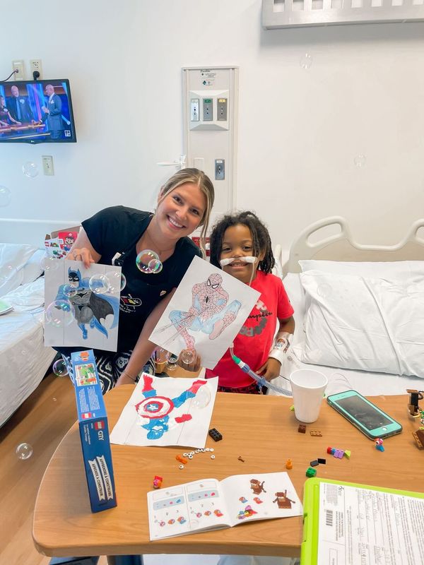 A healthcare worker and a young boy hold up superhero drawings in a hospital room filled with bubbles