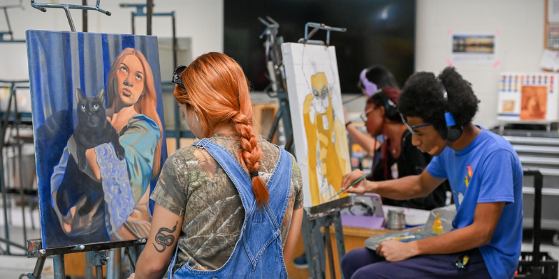 Students sitting at a table showing off their paintings and drawings