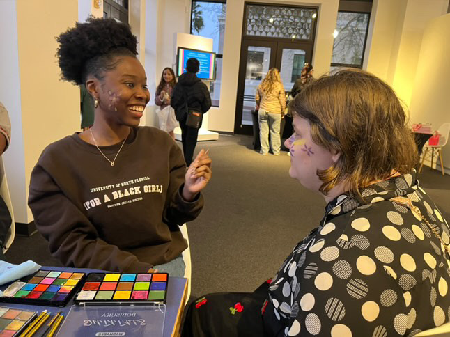 Two women engaged in conversation at a face painting booth at a community event