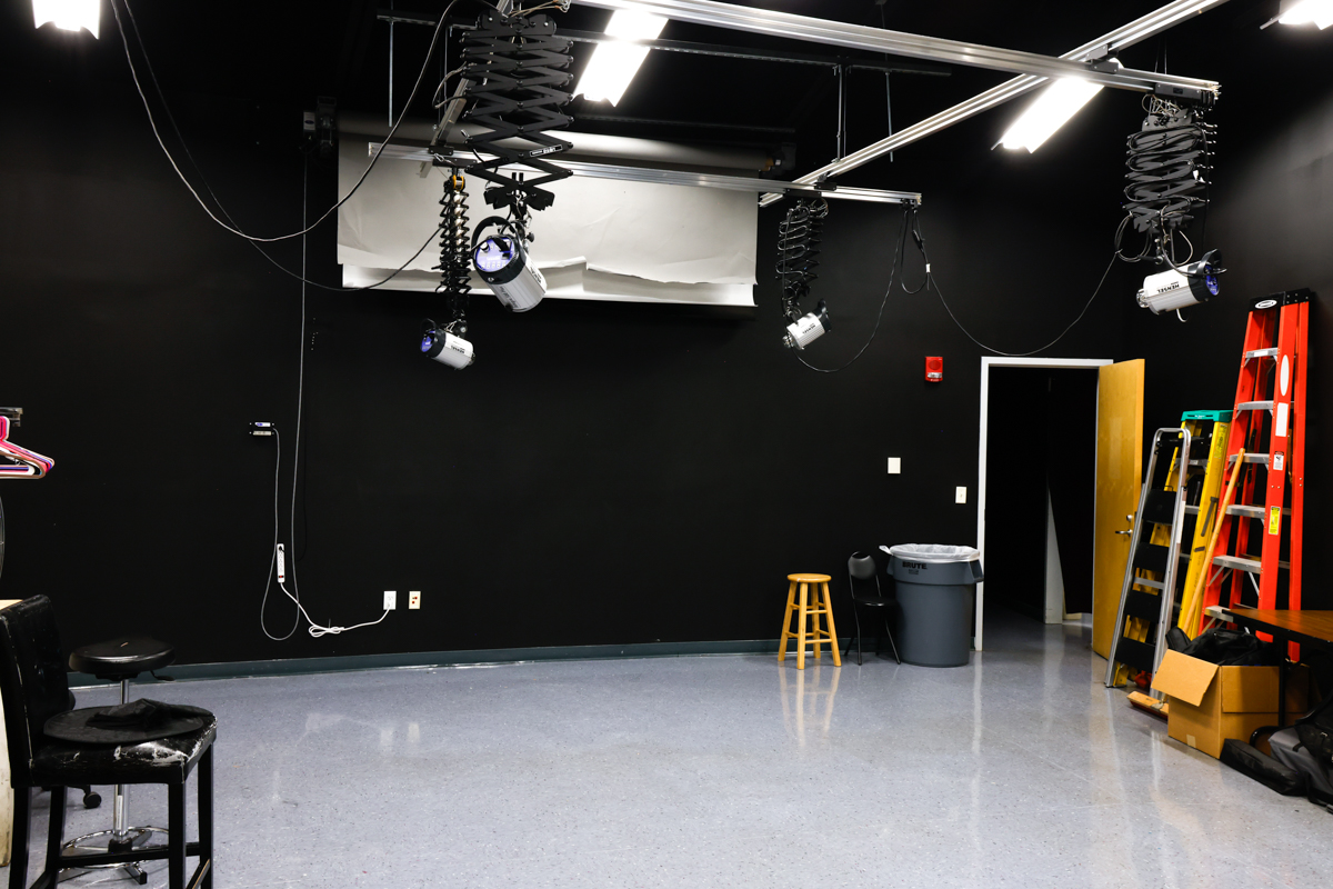 A photography studio with black walls, overhead lighting fixtures, and a partially visible ladder.