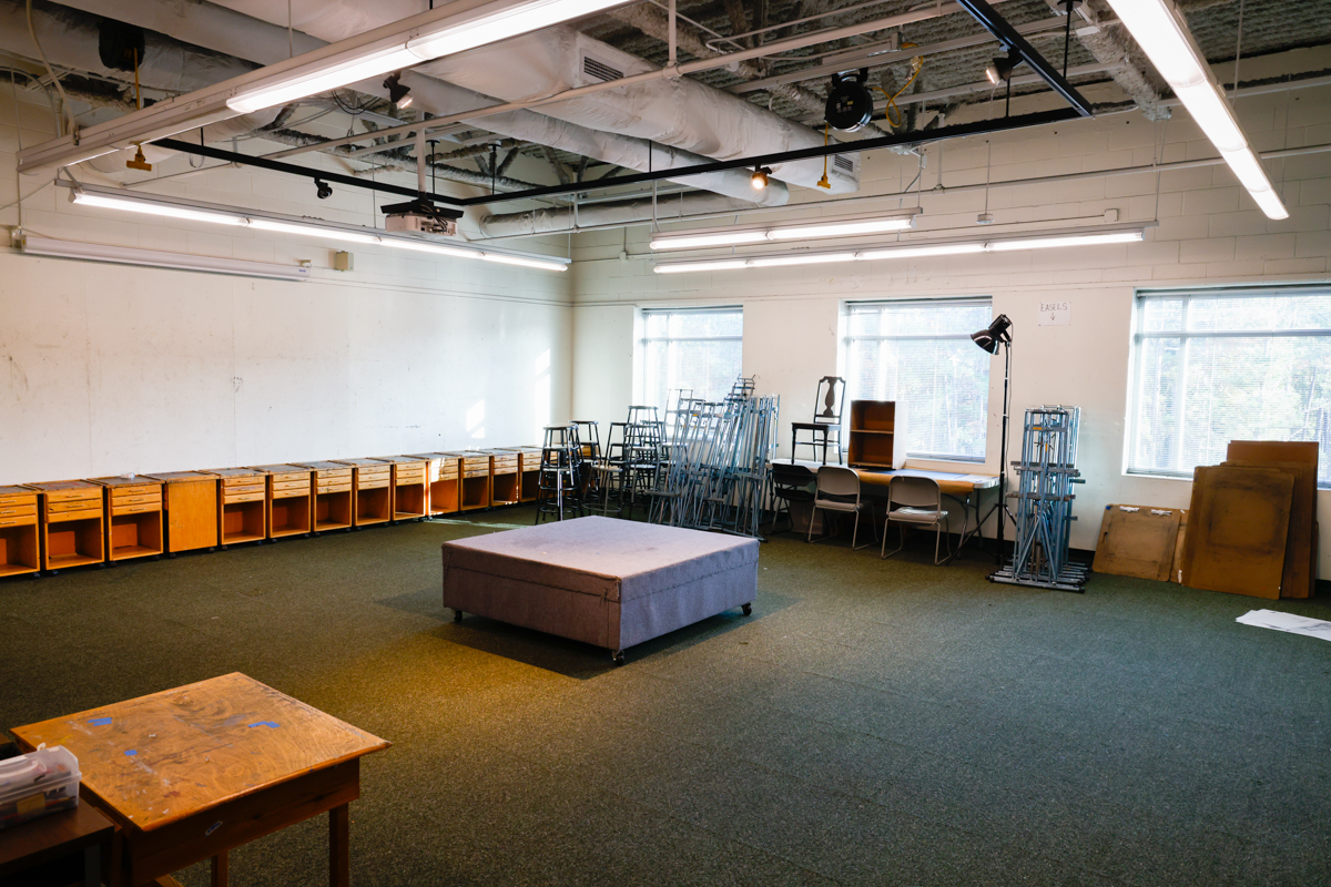 A spacious classroom setting featuring a central gray platform, wooden storage units along one wall, and stacked metal chairs in the background.