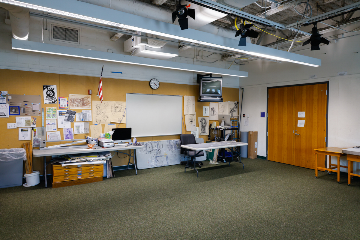 Classroom interior featuring a whiteboard, desk with a computer, and various educational materials on the walls. A wall clock and television are also visible in the space.