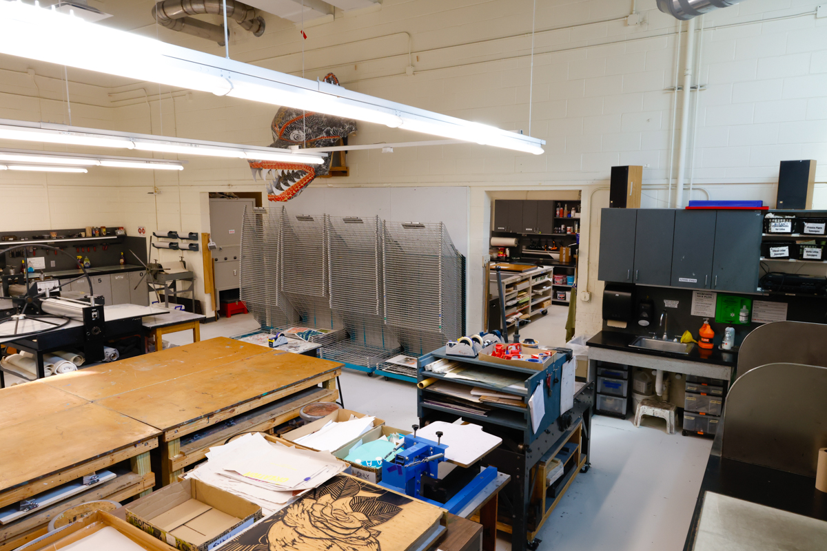 A well-lit workshop featuring various workstations, tools, and organized materials, with a large dinosaur sculpture visible in the background.