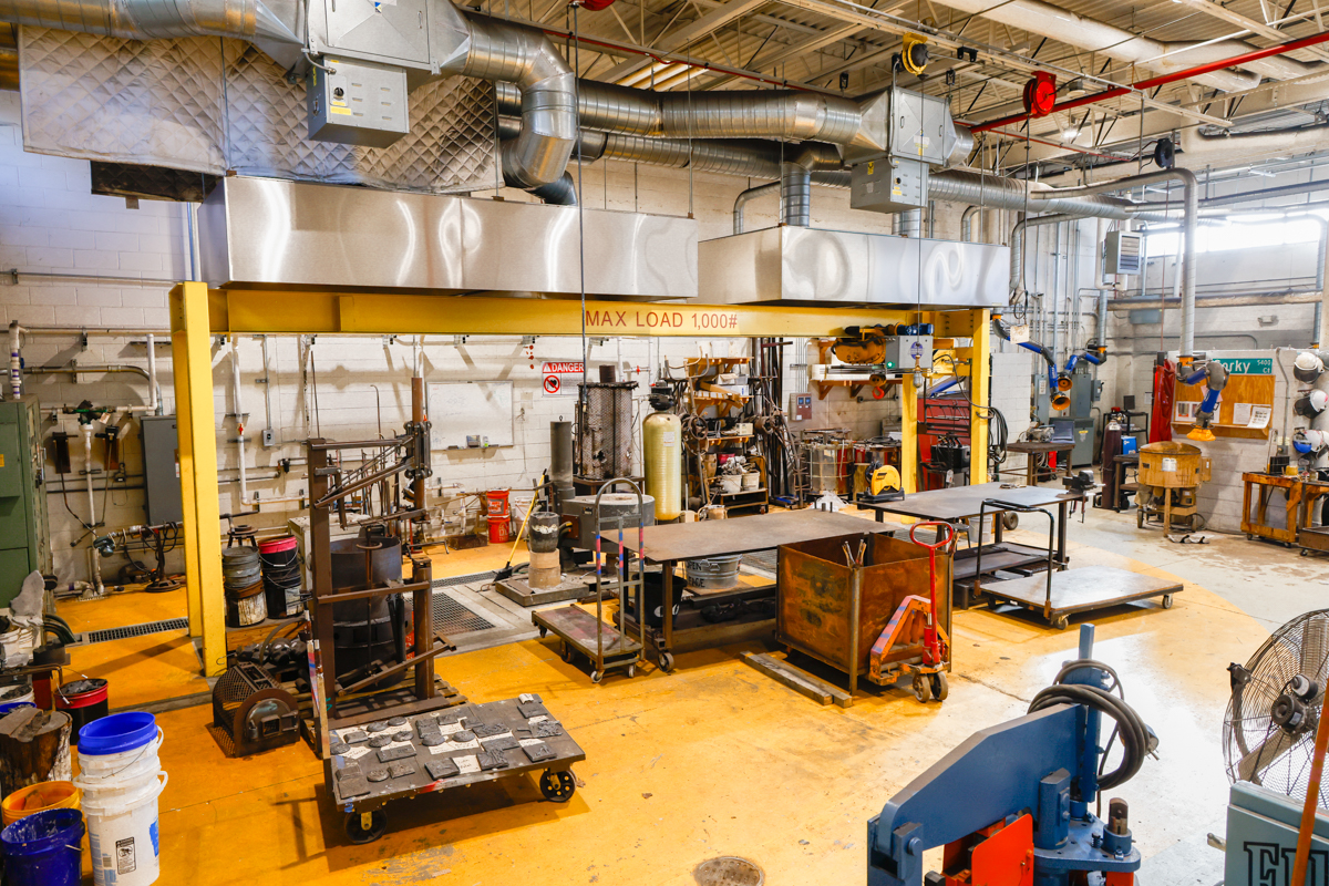 An industrial workshop featuring a large overhead crane, various worktables, and equipment for fabrication and assembly tasks.