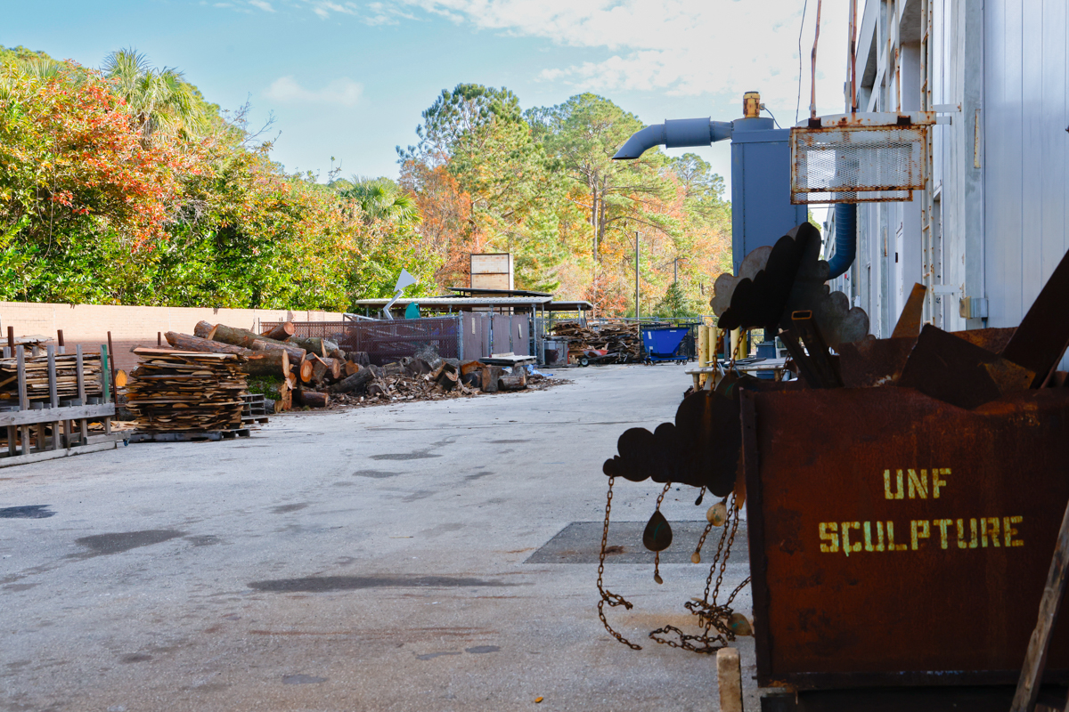 A view of an industrial area featuring a sculpture workshop with wooden logs stacked on one side and a rusty container labeled "UNF Sculpture" on the other.