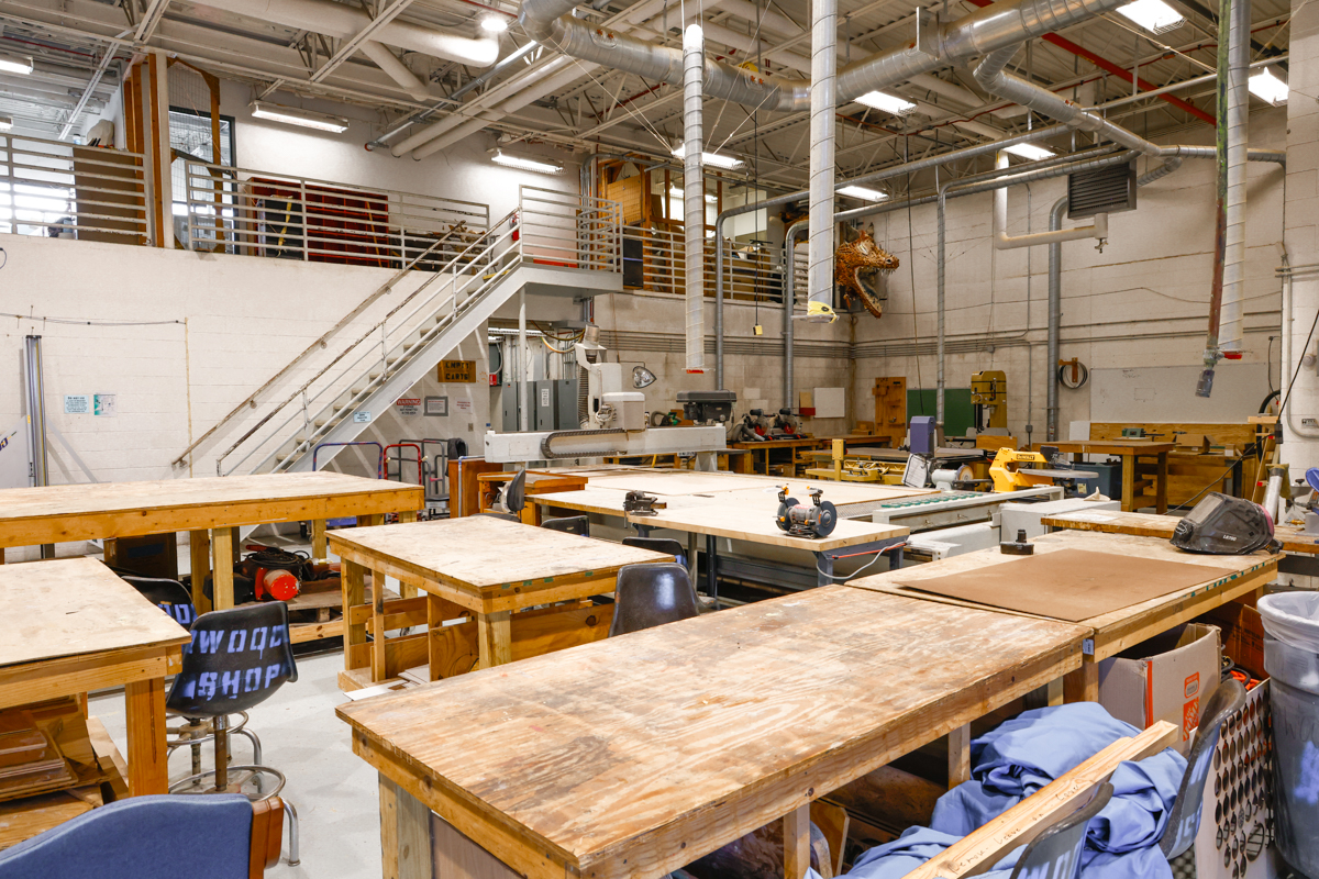 Interior view of a woodworking shop featuring various worktables, tools, and equipment. The space has a spacious layout with upper-level access and ample natural light.