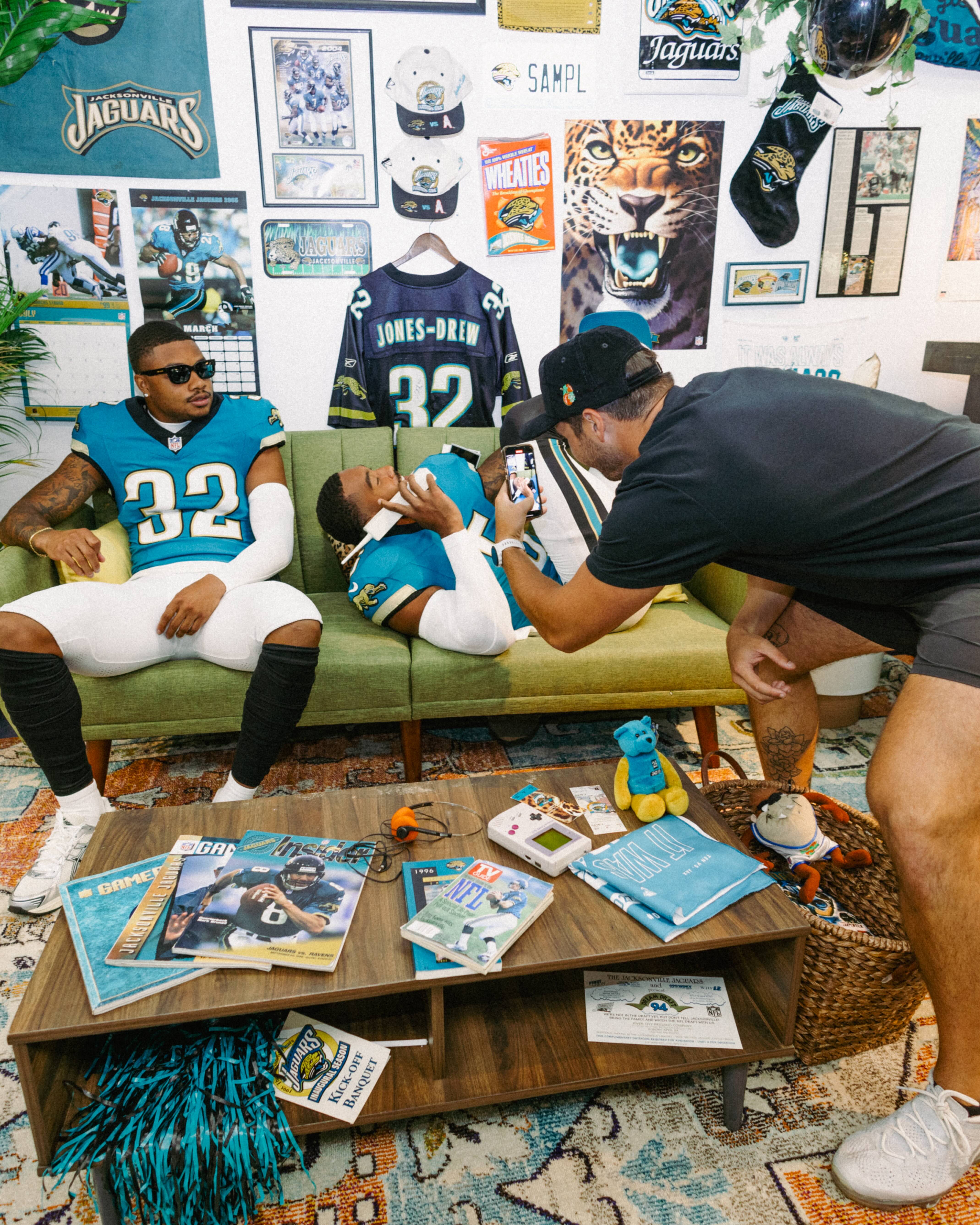 Two football players in Jacksonville Jaguars uniforms sit on a couch while a third person takes their photo, surrounded by sports memorabilia and decor.