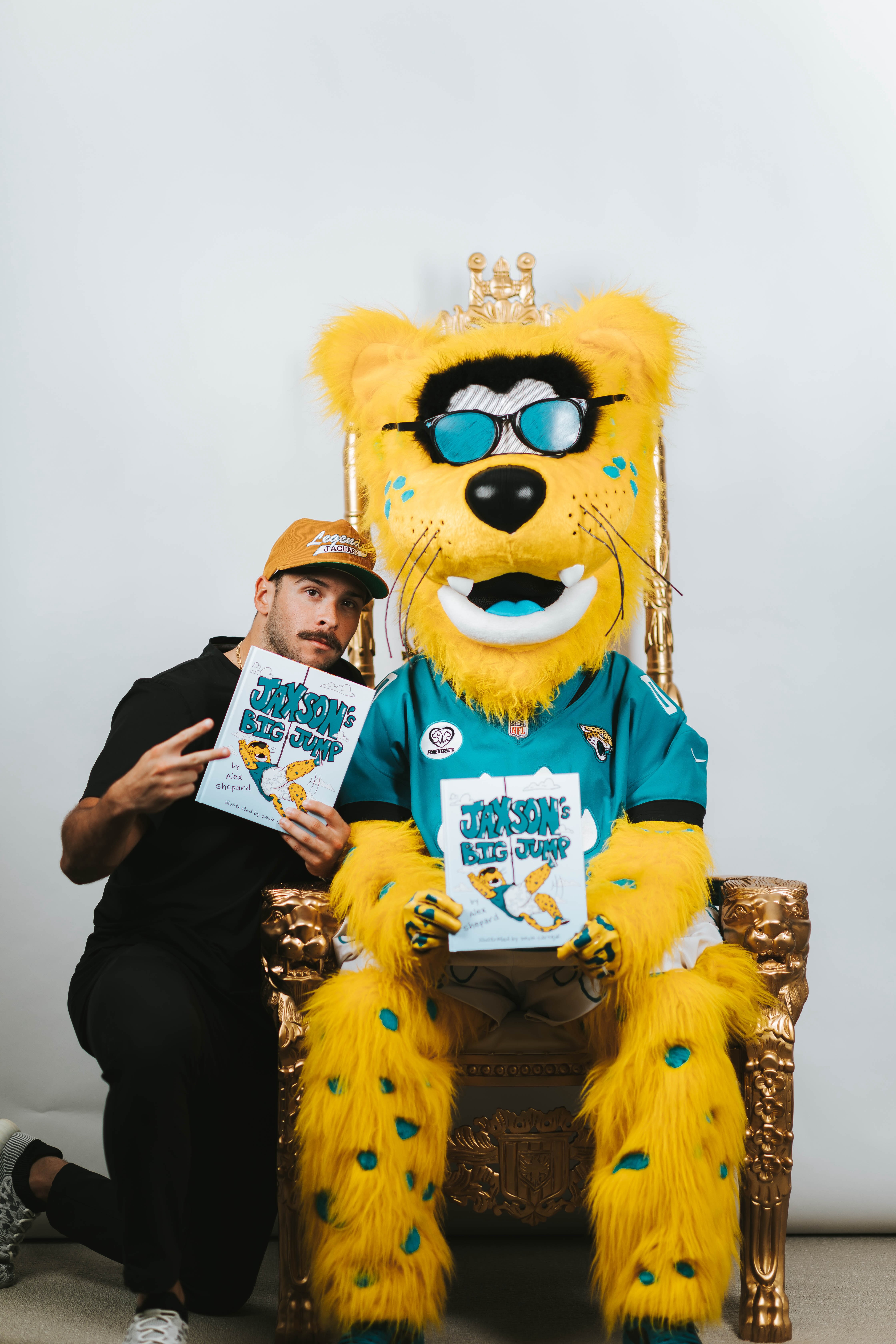 A person posing next to a large, yellow cartoon mascot dressed in a sports jersey, holding a children's book titled "Jaxson's Big Jump."