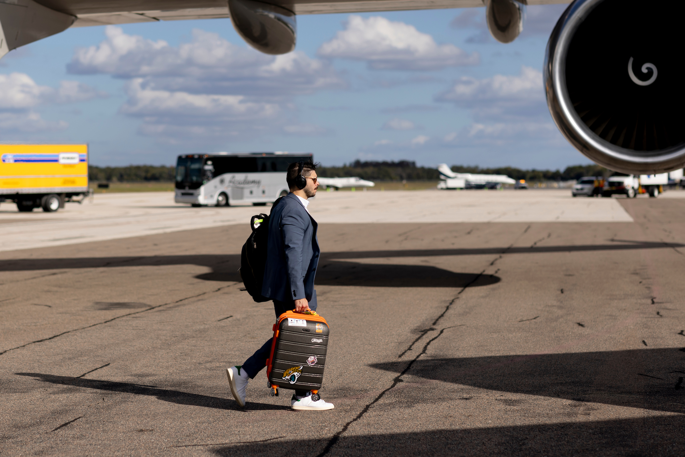 A man in a suit walks with a suitcase at an airport, with aircraft and a bus in the background.