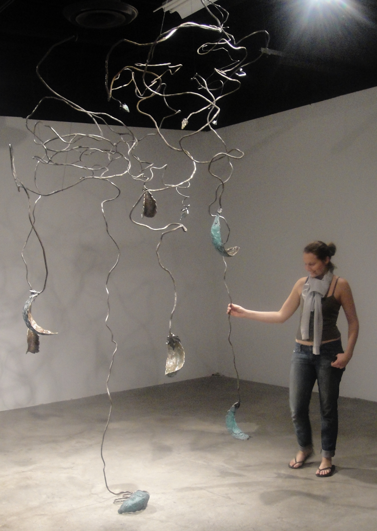 A woman interacts with a modern metal mobile sculpture featuring abstract twisted shapes and hanging elements in a gallery setting.