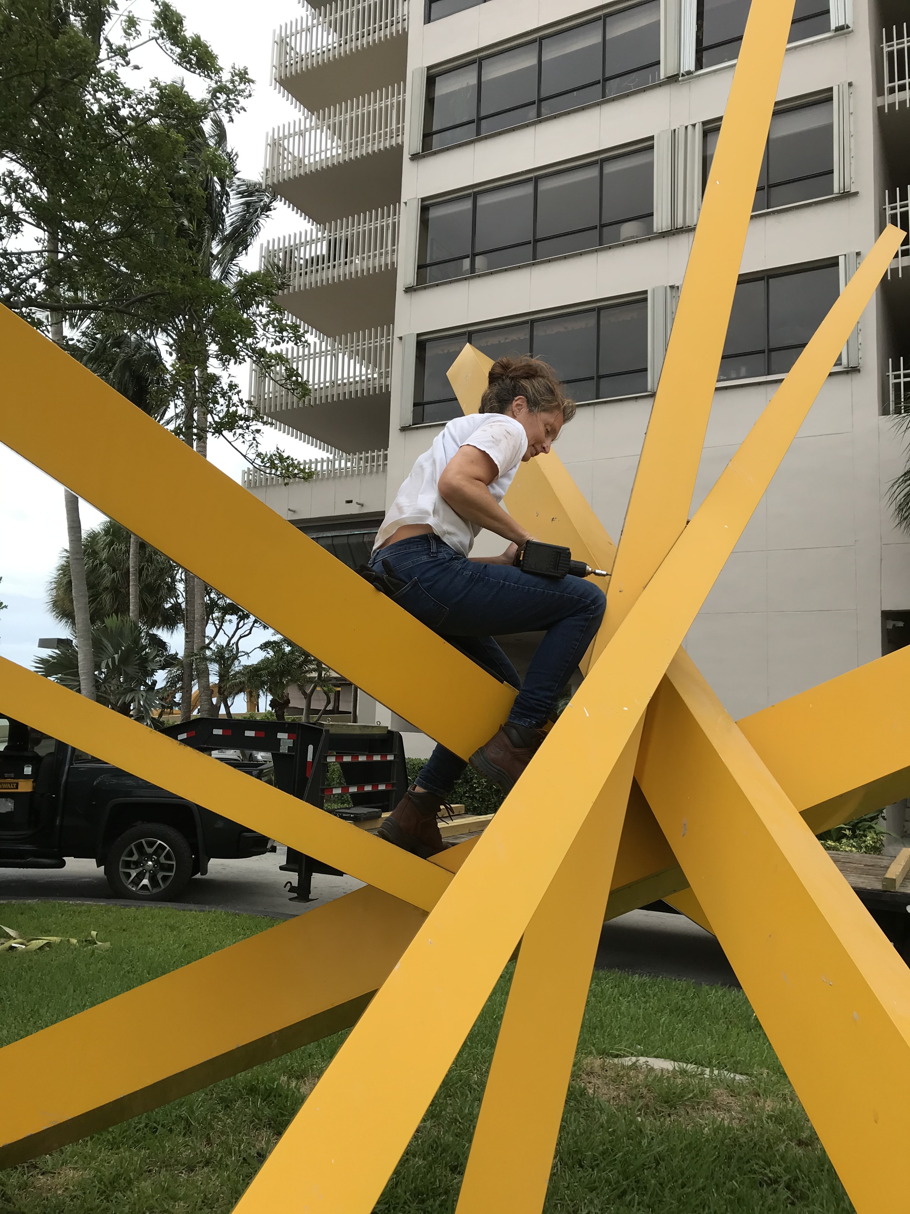 A person climbing on a large yellow outdoor sculpture with star-like beams near a modern building.