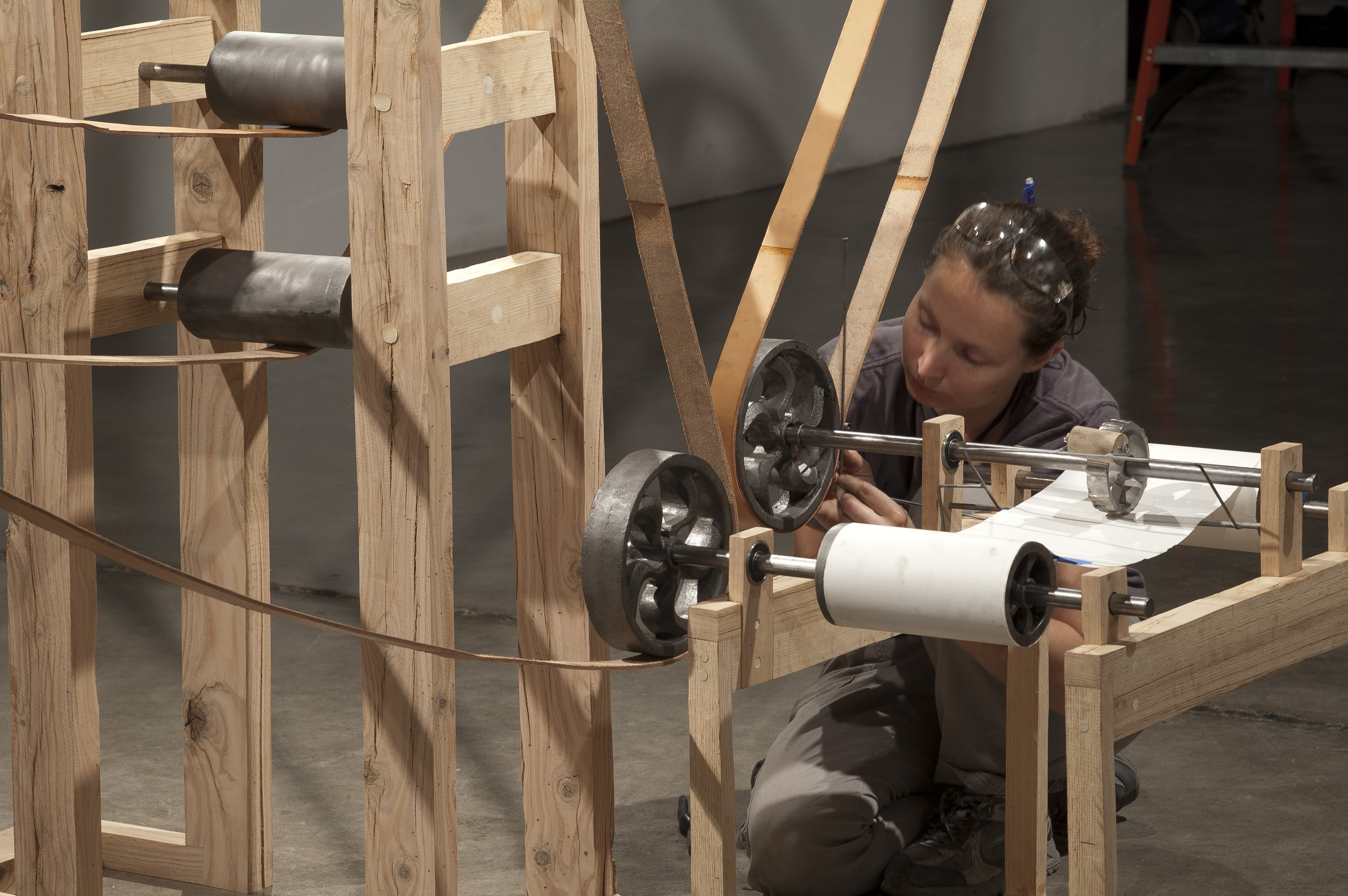 A woman works on a wooden kinetic sculpture, adjusting its mechanisms and rolls. The image showcases the intricate details of the artwork and the artist's engagement with the piece.