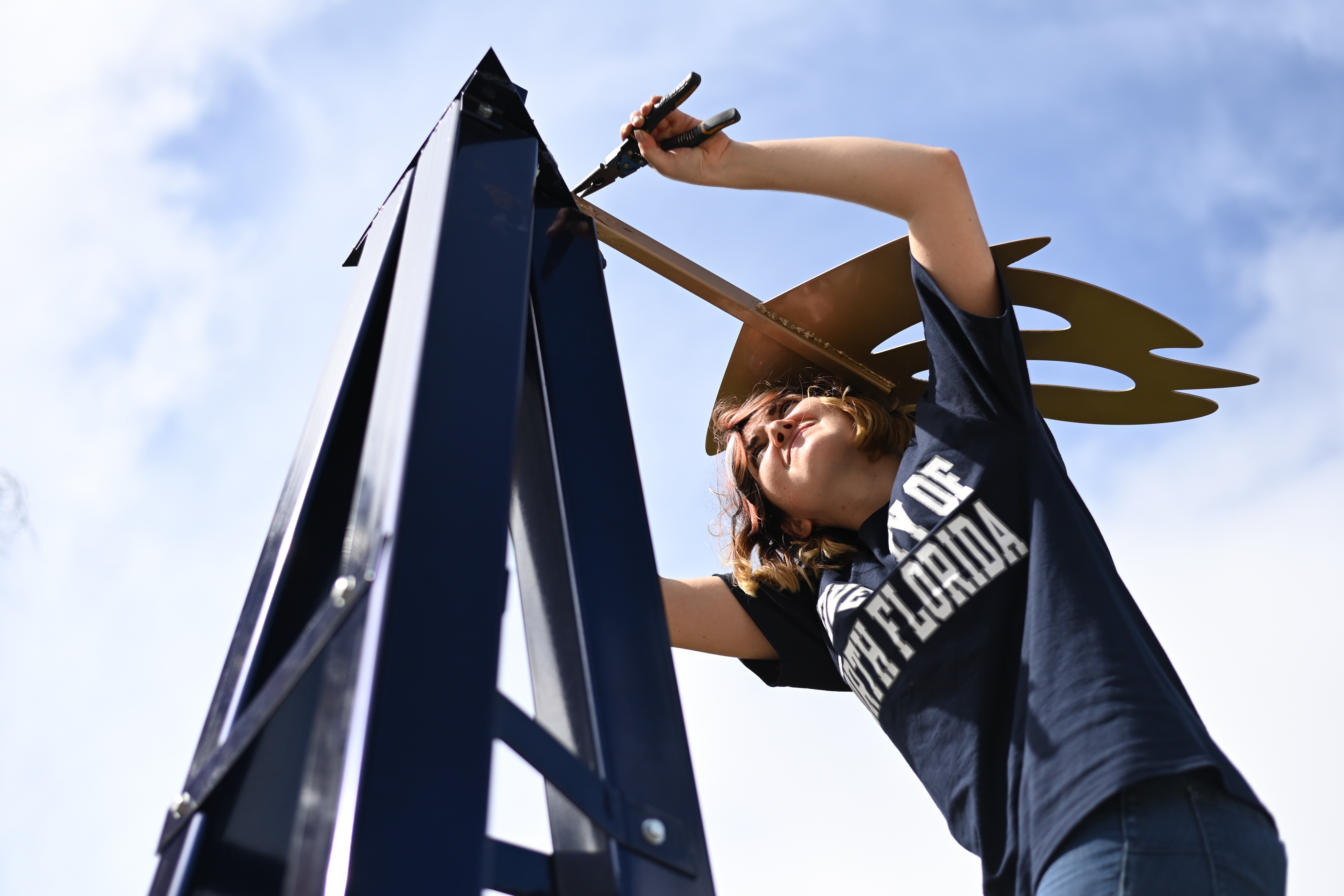 A young person adjusting a large, golden wind turbine blade on a metal structure against a blue sky.