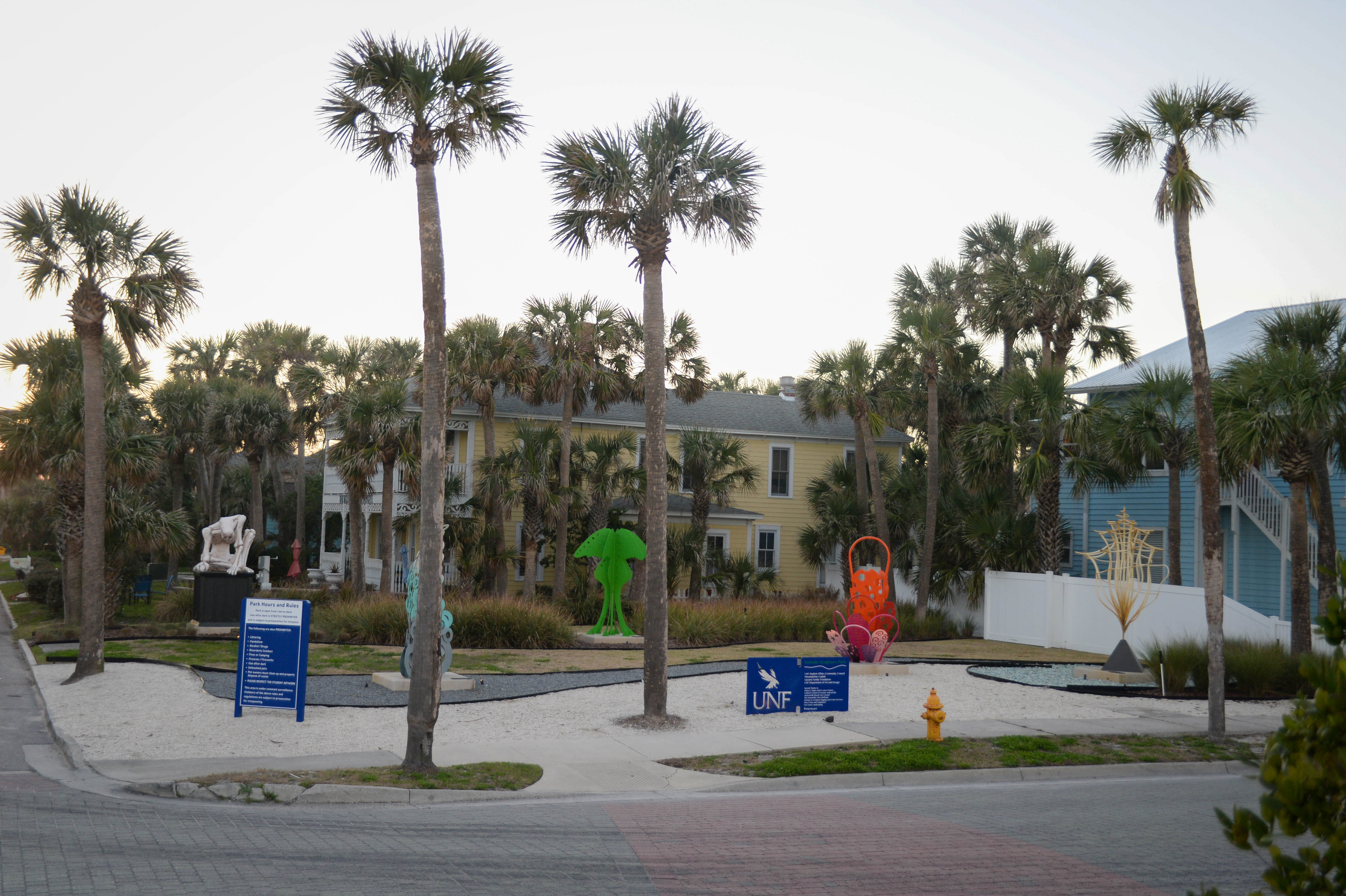 A coastal park scene featuring palm trees, various sculptures, and a sign for the University of North Florida (UNF).