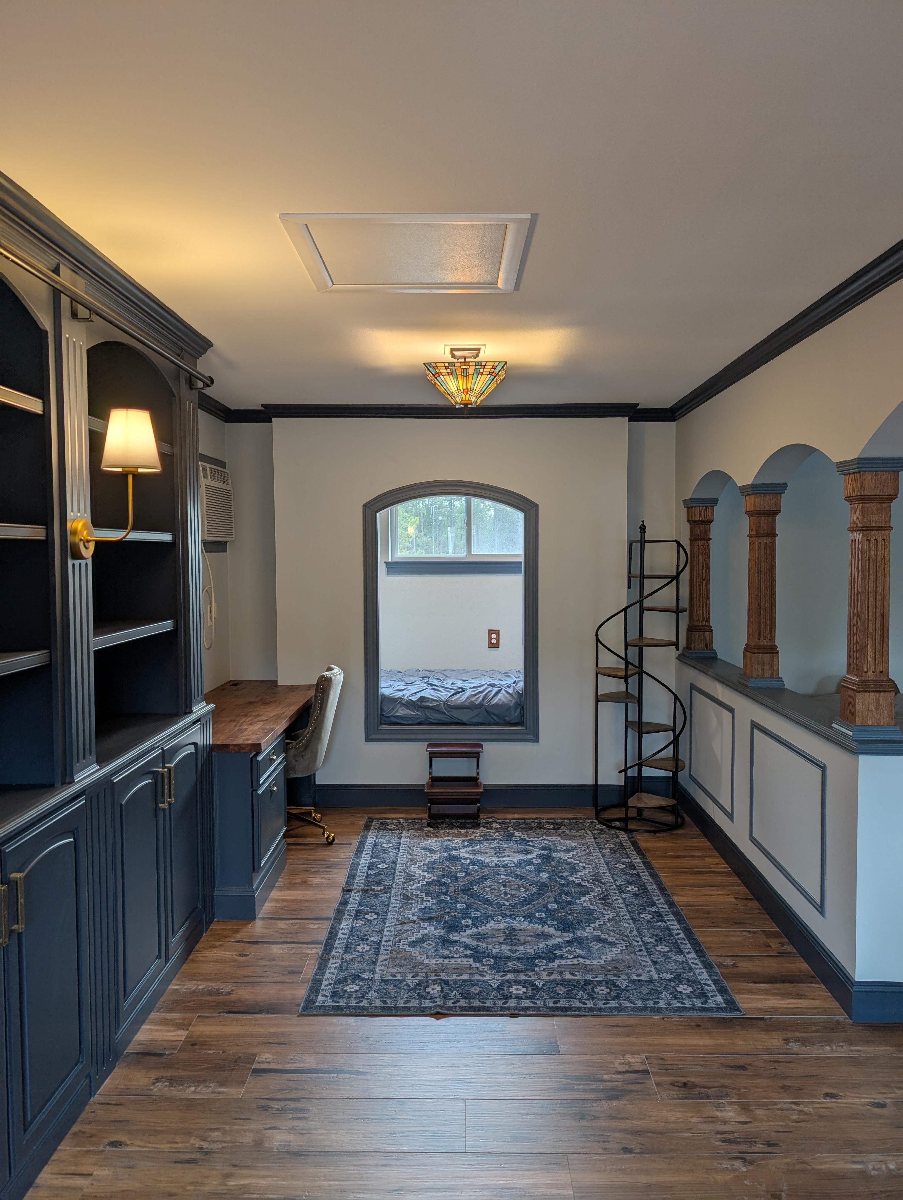 A cozy, modern workspace featuring a dark blue built-in bookshelf, a stylish spiral staircase, and a decorative rug on wooden flooring.