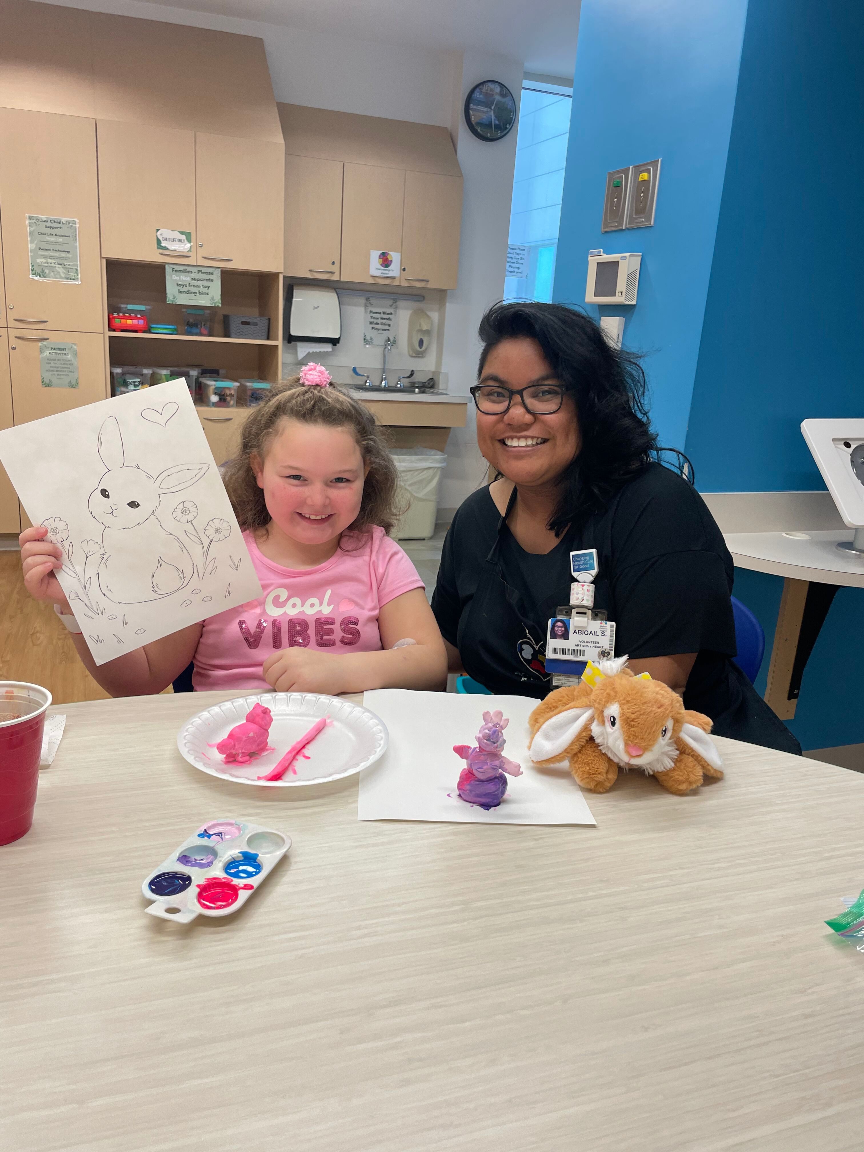 A young girl and a caregiver smile while creating colorful clay bunnies at a table, with a drawing of a bunny in front of them.