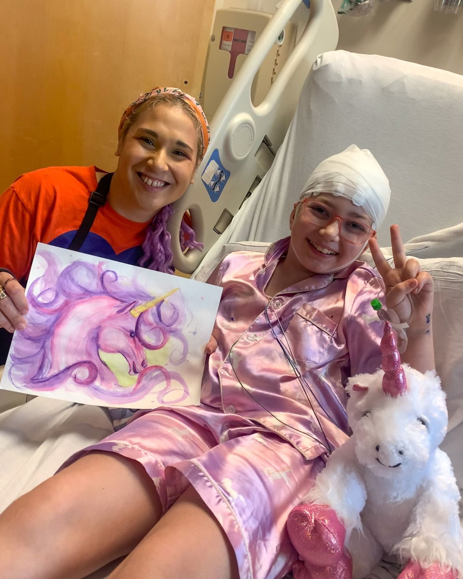 A patient in a hospital bed smiles while holding a colorful unicorn drawing, accompanied by a caregiver and a plush unicorn toy.