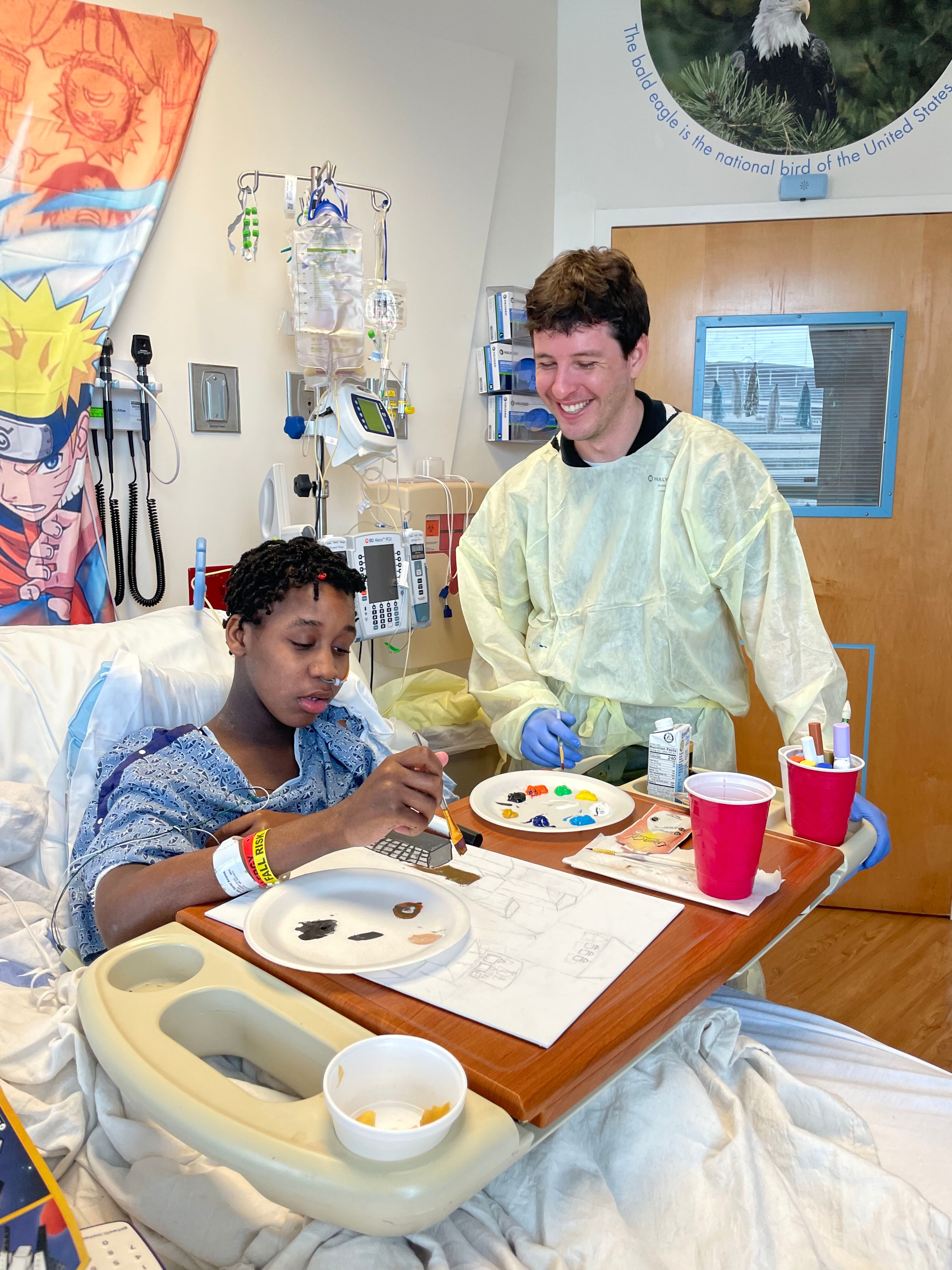 A healthcare volunteer engages with a young patient, who is painting, in a hospital room decorated with colorful artwork.