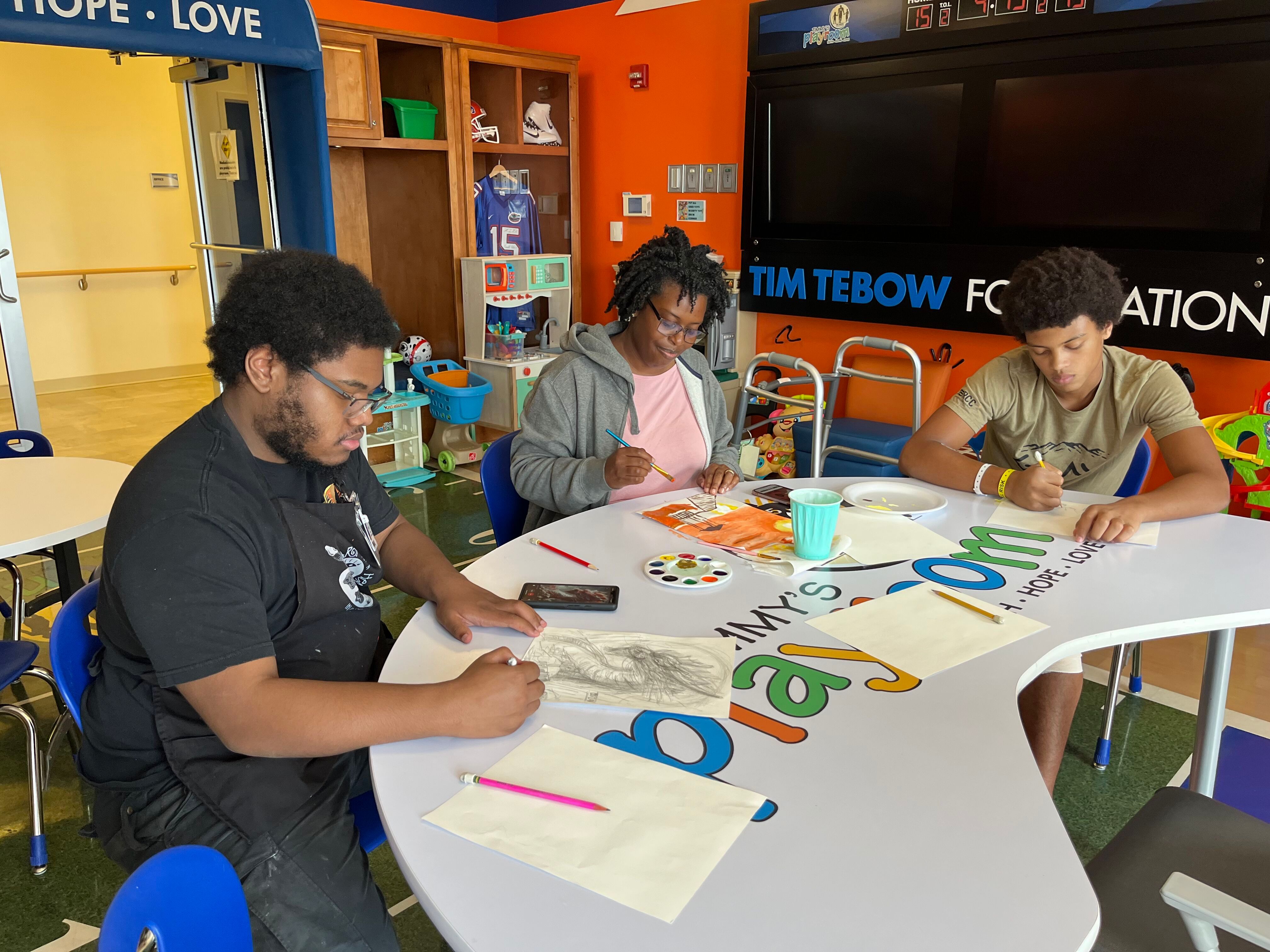 Three young individuals engaged in an art activity at a colorful table in a creative play space, each focused on their drawings.