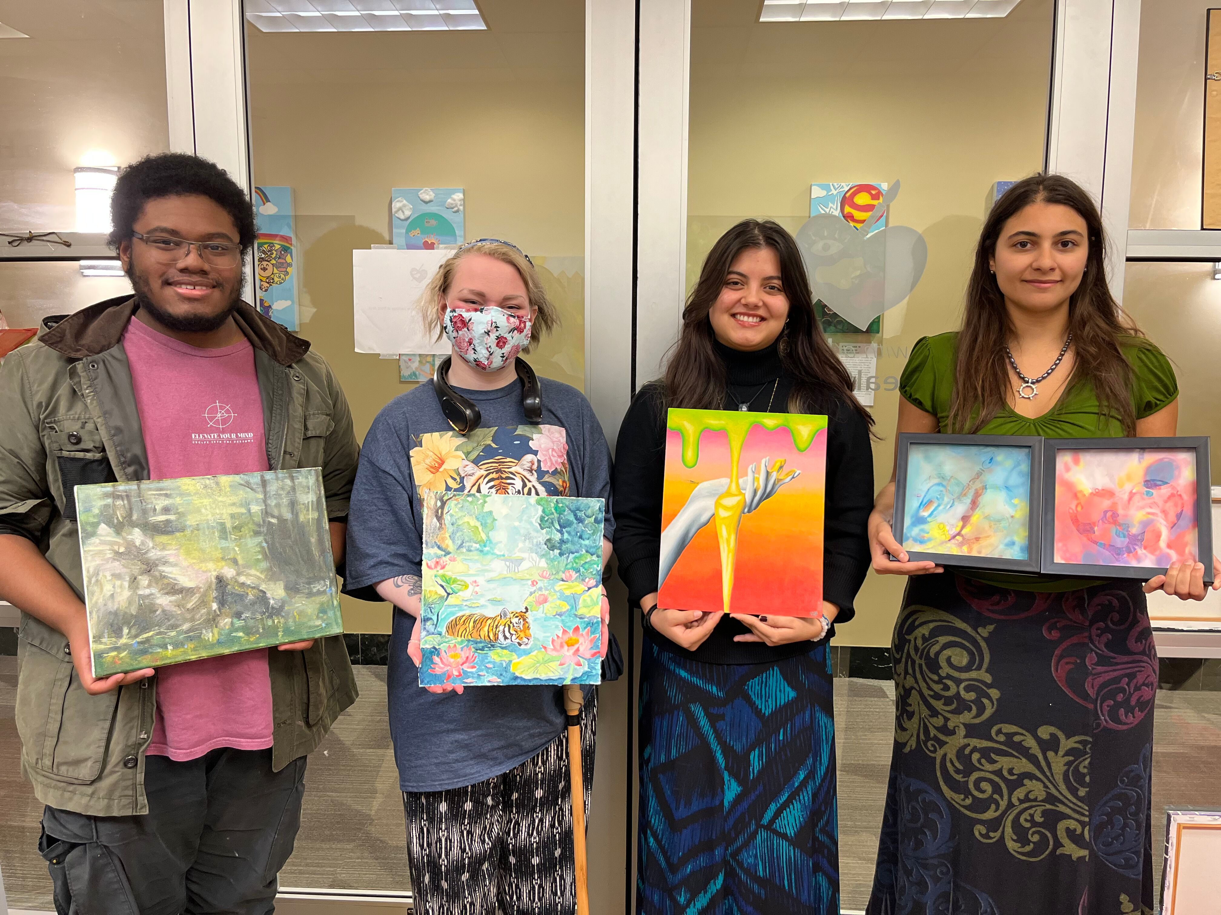 Four art students proudly display their paintings in a classroom setting.