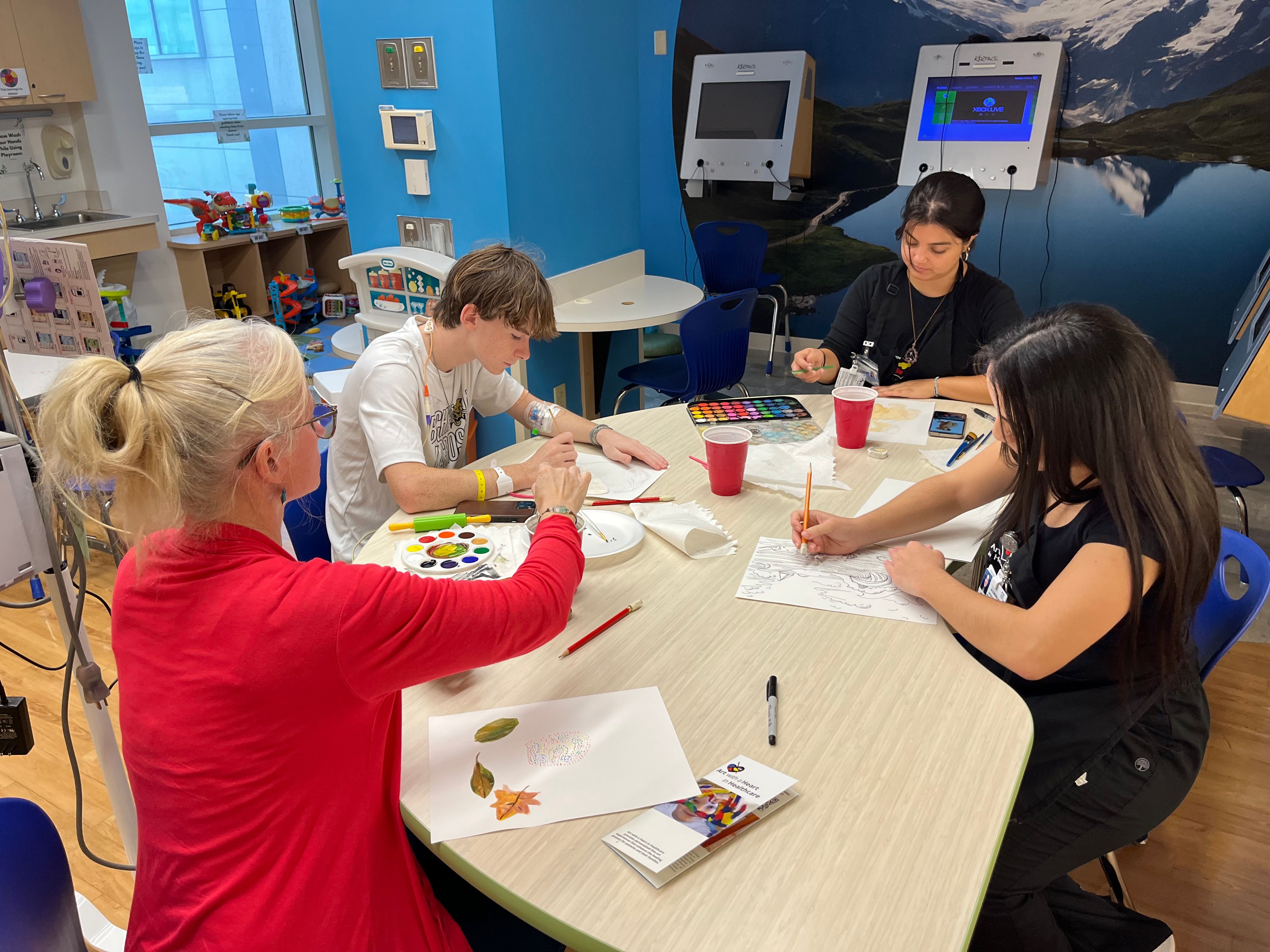 A group of four individuals engaged in a creative art session at a colorful table, using paints and colored pencils. The setting appears to be a bright and inviting space, likely a hospital or healthcare facility.