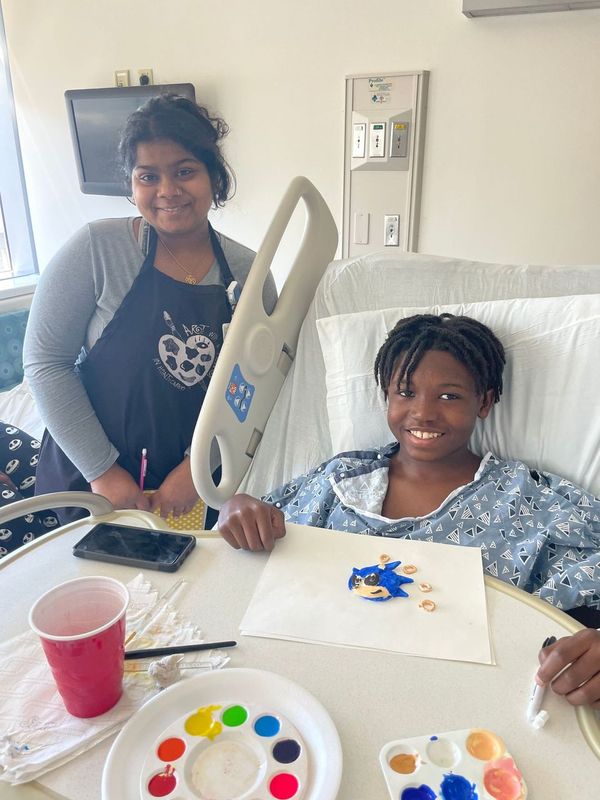 A patient in a hospital bed smiles while holding a painted Sonic the Hedgehog, accompanied by a caregiver.