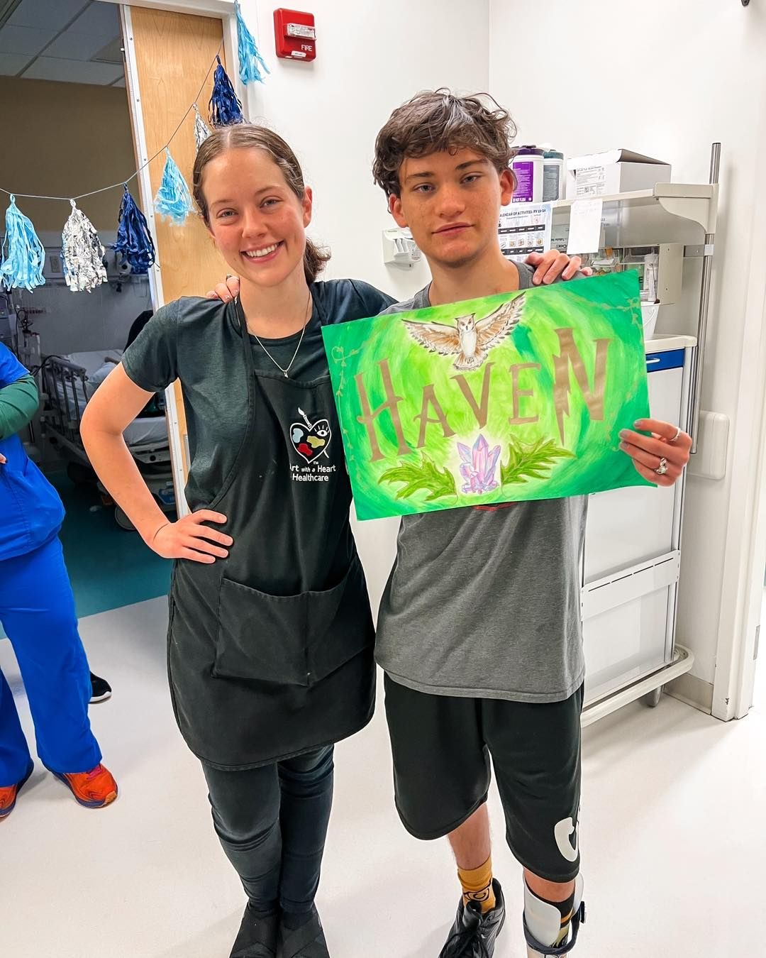 A young man and a woman stand together, smiling while holding a colorful sign that reads "HAVEN" in a hospital setting.