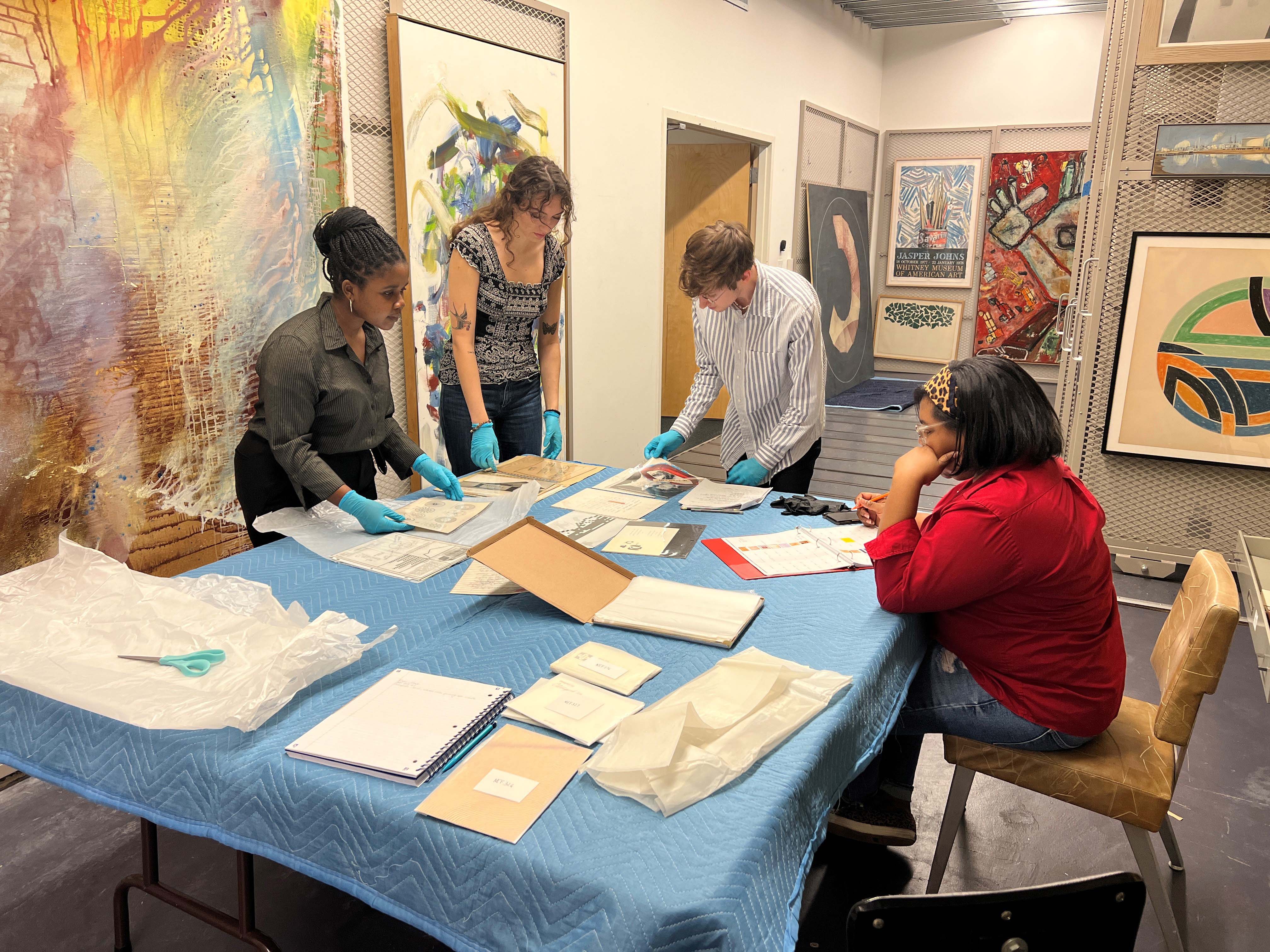 A group of four people wearing gloves is examining artwork and documents on a blue table in an art studio.