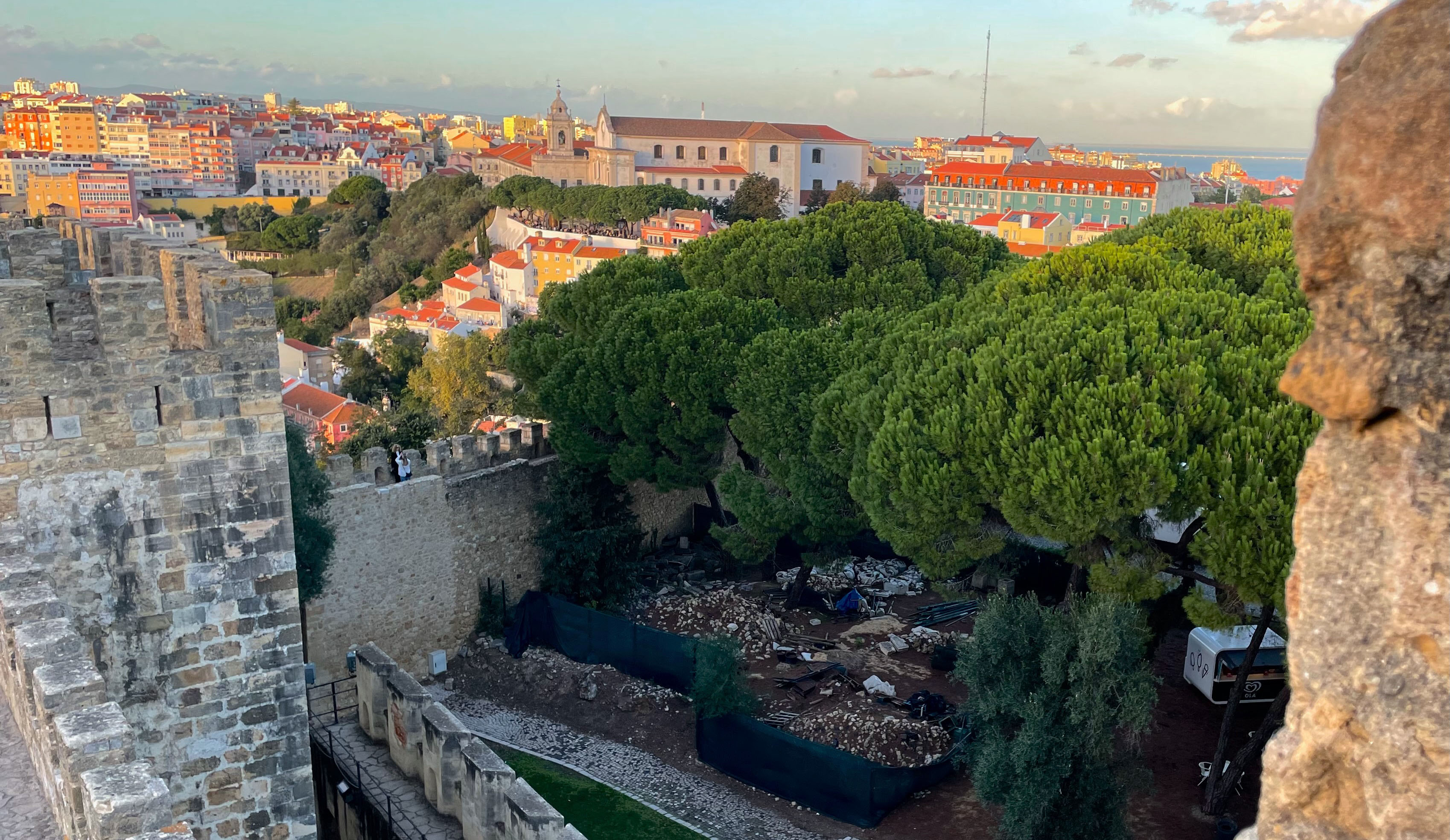 A panoramic view of the castle of São Jorge featuring a castle wall with lush green trees and colorful buildings in the background.