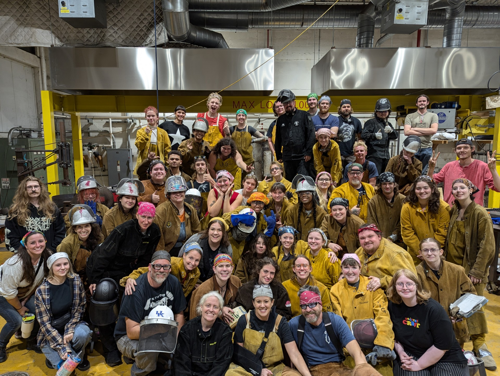 A large diverse group of people wearing welding gear poses together in a workshop setting, showcasing camaraderie and teamwork.
