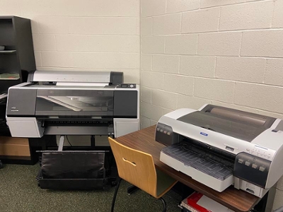 Two large office printers positioned next to each other in a workspace, with a wooden chair in front of one.