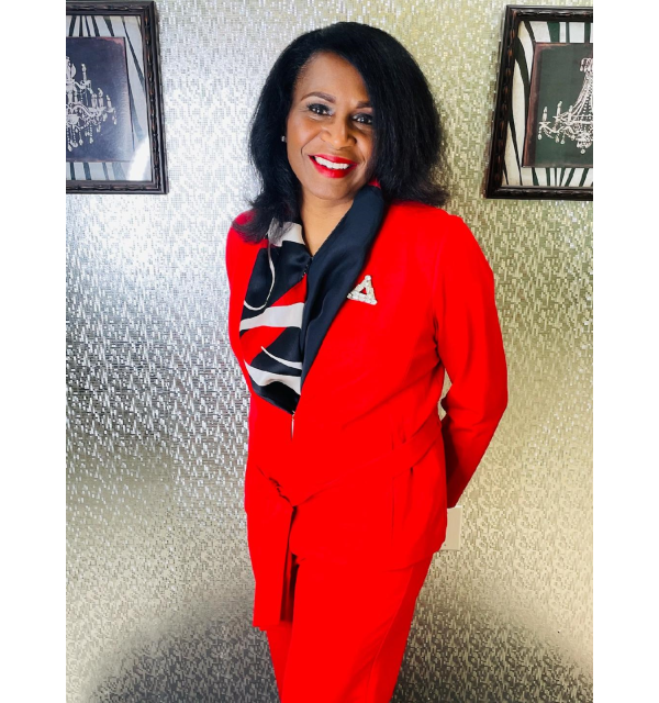 Dr. Perkins in a red suit with a patterned wall and two hanging pictures behind her.
