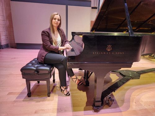 Woman with long blond hair and burgundy blazer sitting next to piano.