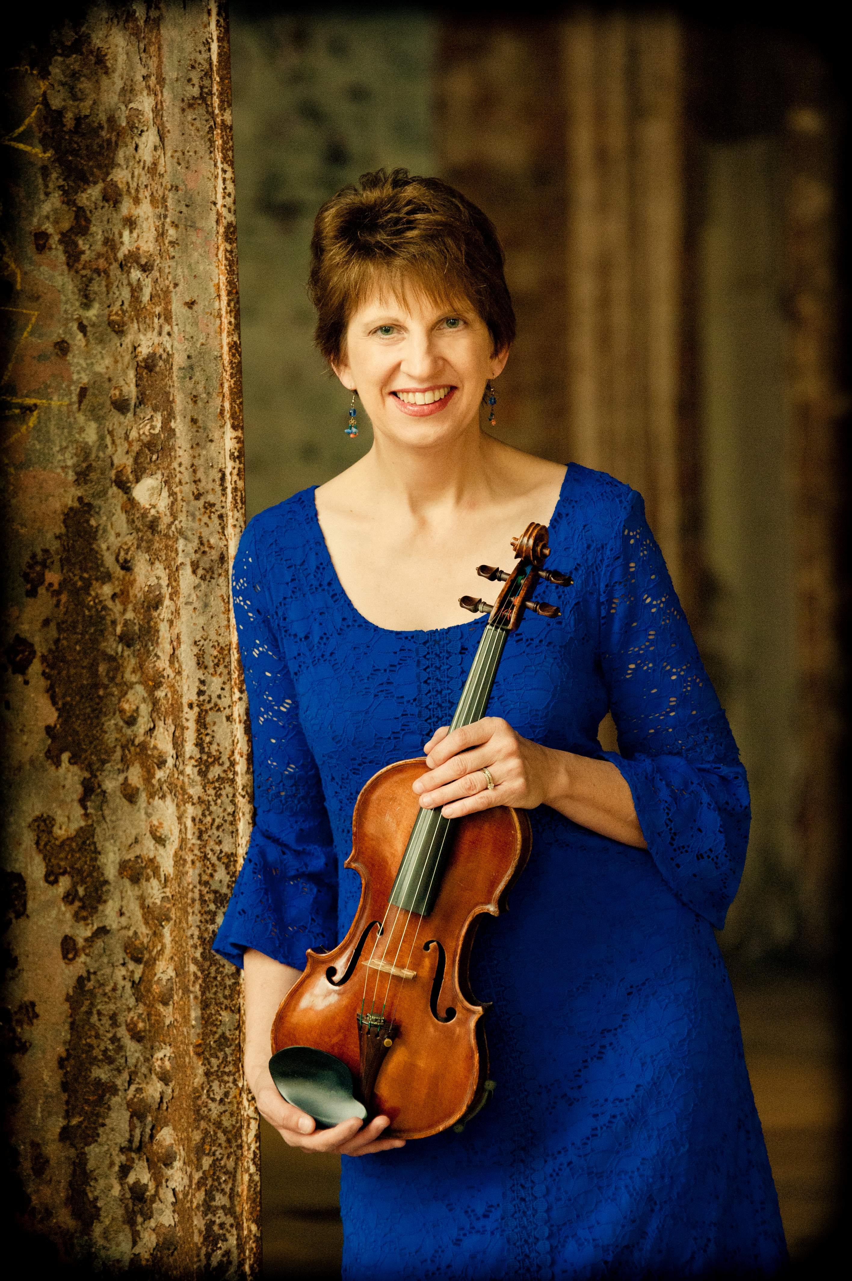Woman with short brown hair wearing a long blue dress and holding a violin. 