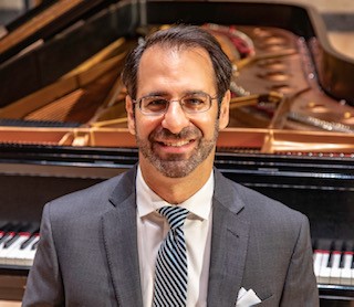 Man sitting in front of piano in gray suit with striped tie and wearing glasses. 