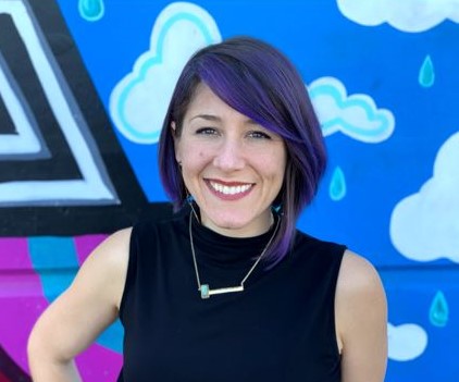 Woman standing in front of colorful wall wearing black short-sleeved top. Her hair is a short bob with purple streaks. 