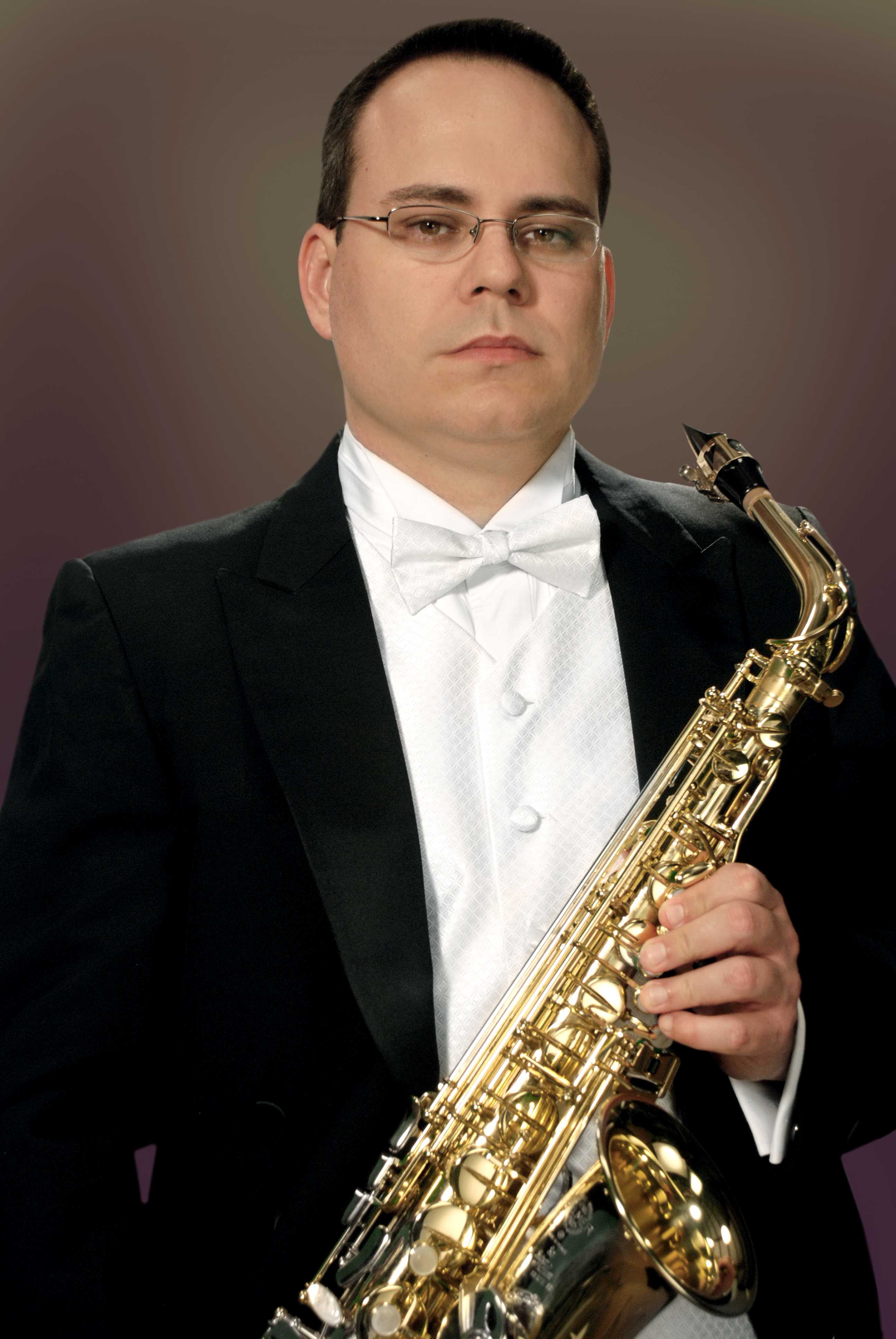 Caucasian male with close cropped black hair wearing a tuxedo with white bow tie. He wears glasses and holds a saxophone. 