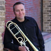 Man in black long-sleeved shirt leaning on ledge and holding a trombone.