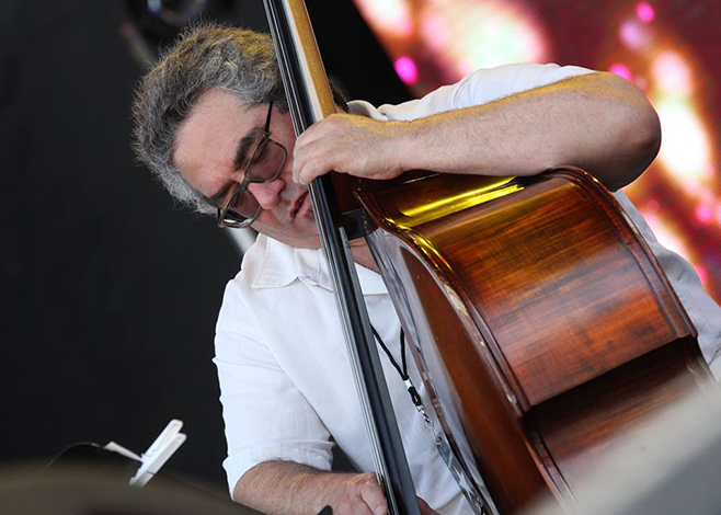 Man with short hair and glasses, playing double bass on stage.