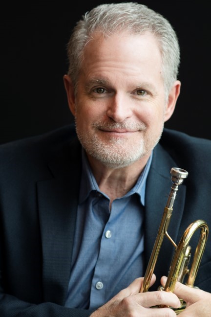 Older gentleman with gray hair wearing a blue blazer and blue shirt and holding a trumpet. 